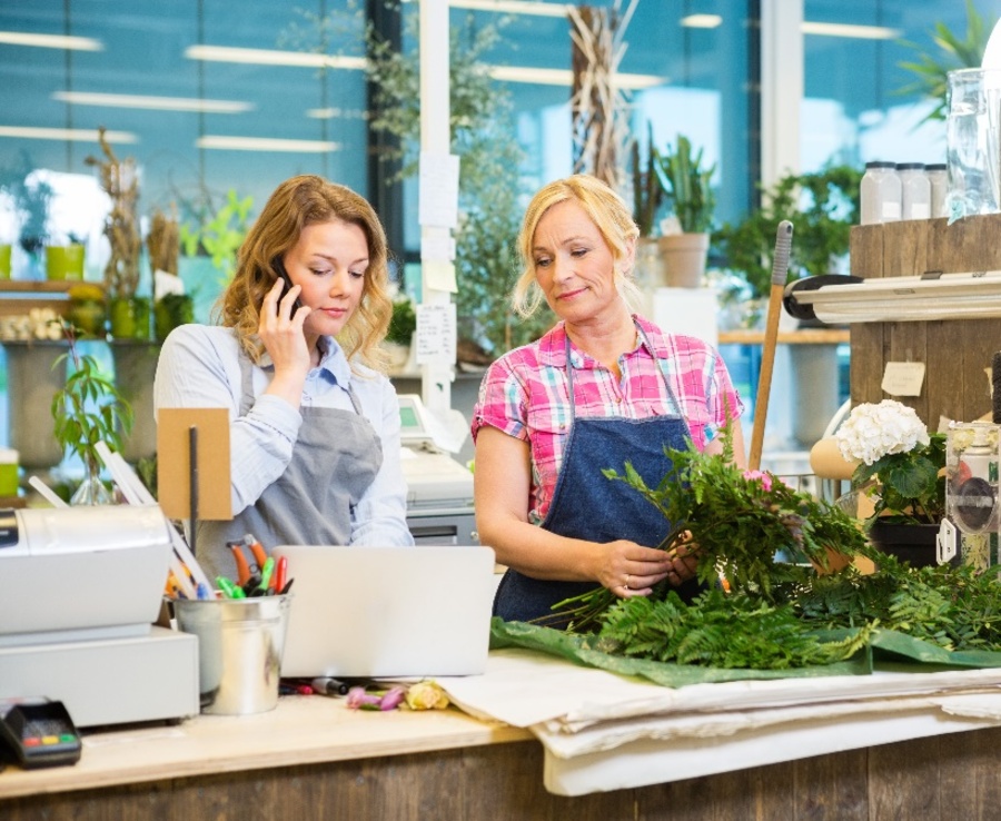 Dve ženy v zástere za kvetinárskym pultom: jedna telefonuje a druhá viaže kyticu