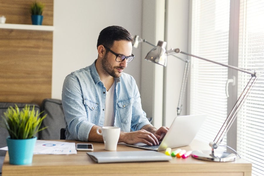 Ung man sitter hemma vid skrivbordet och arbetar på en bärbar dator.