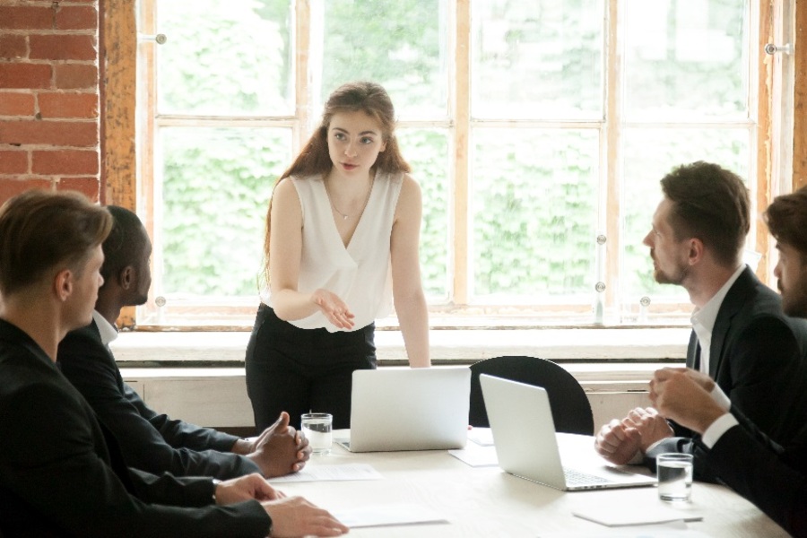 Fiatal nő, aki egy tárgyalóteremben férfi beosztottjaihoz beszél