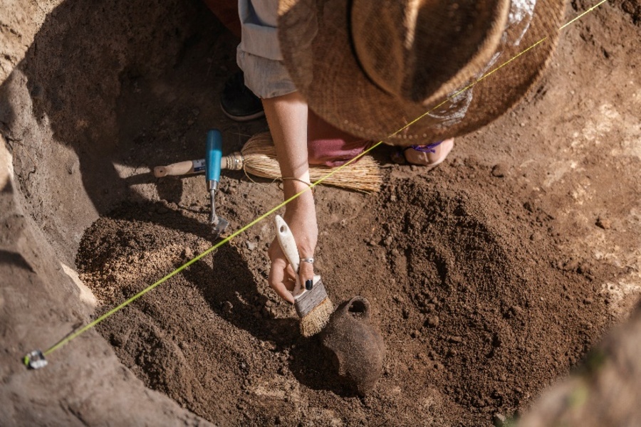  Sun hat-wearing archaeologist squatting in an excavation site, uncovering an old ceramic object with a brush