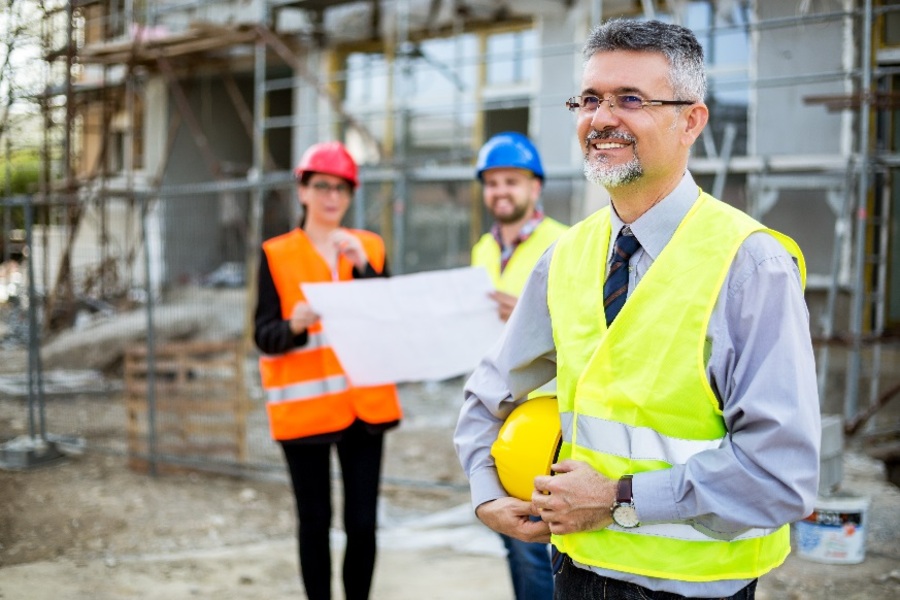 Três operários de construção num estaleiro com coletes de proteção, dois de pé, ao fundo, com capacetes de proteção, a segurar num projeto.