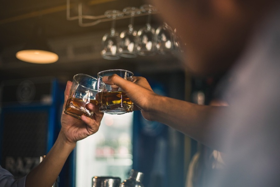 Two people's hands clinking glasses filled with caramel-coloured spirits in a bar
