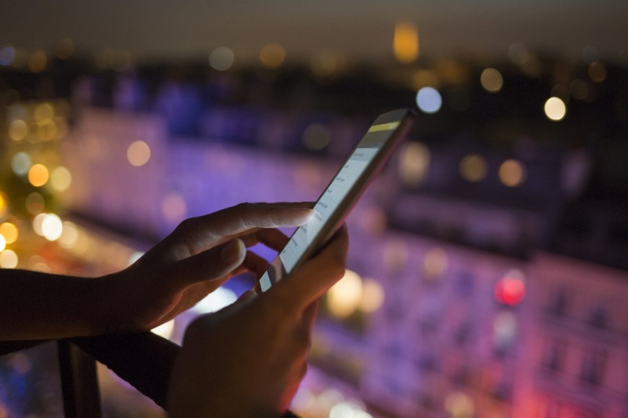 Hands holding a mobile phone on a balcony at night against a backdrop of sparkling city lights