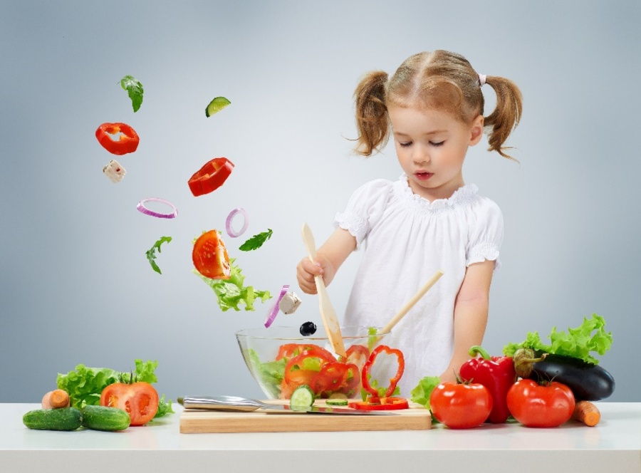  Young girl stirring sliced vegetables in a bowl, various whole vegetables to both sides of her, while cut vegetables fly out of the bowl and float in the air