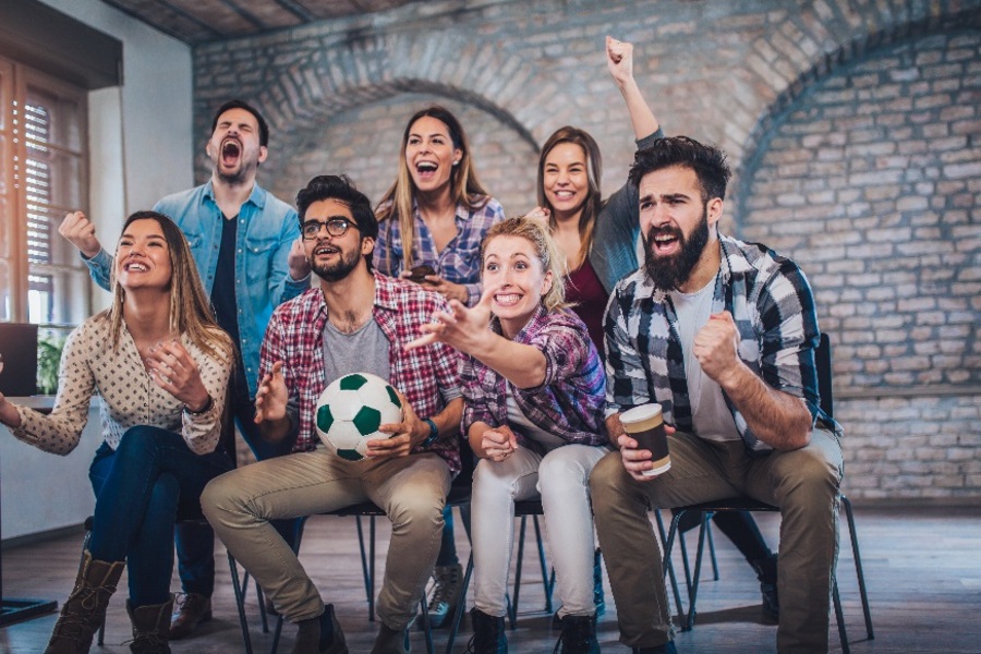 Zeven enthousiaste voetbalfans, van wie er één een voetbal vasthoudt, kijken in een overdekte ruimte samen naar een wedstrijd