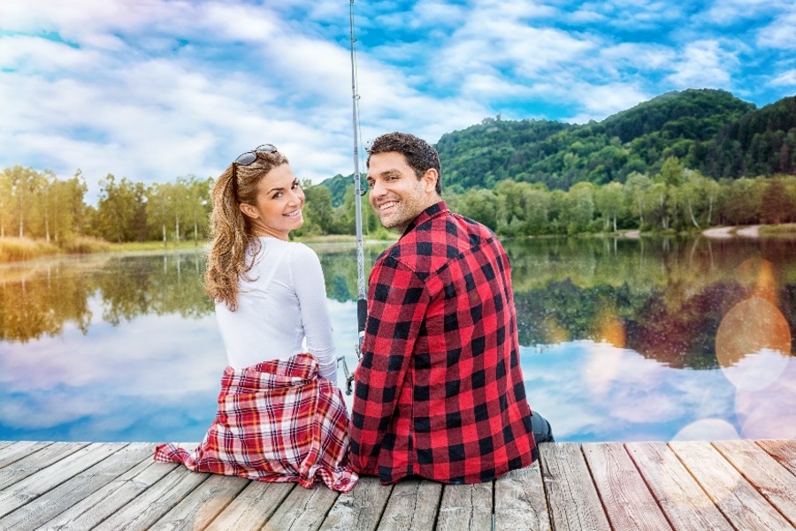 Para młodych ludzi siedzi z wędką na pomoście jeziora. Odwracają się do kamery i uśmiechają się.