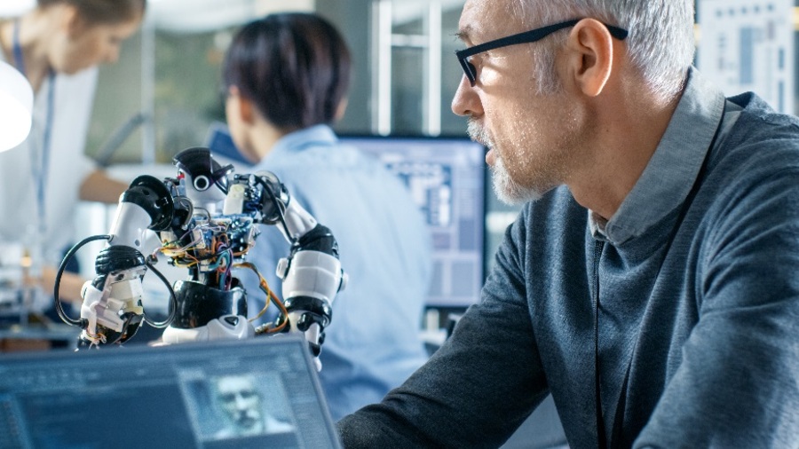 Člověk v laboratorním prostředí manipuluje s robotem vedle obrazovky notebooku, na které je zobrazen program pro rozpoznávání řeči a obličeje