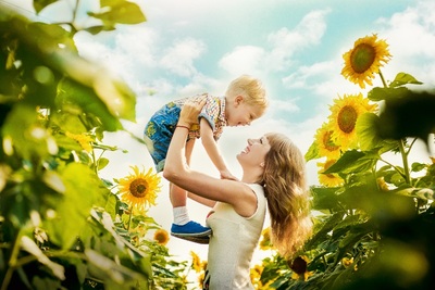 Frau, die ihren jungen Sohn in einem Sonnenblumenfeld hochhält, im Hintergrund ist ein blauer Himmel mit kleinen Wolken zu sehen