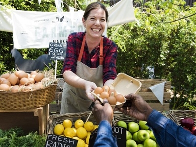 Női mezőgazdasági termelő egy doboz biotojást ad át széles mosollyal egy vevőnek a helyi piacon