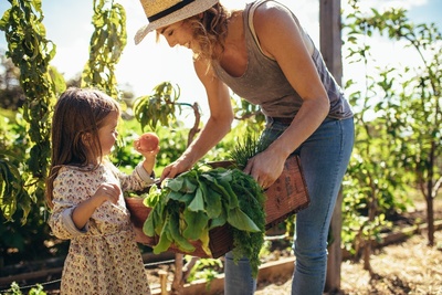 Eine junge Frau hält eine Kiste mit frischem Obst und Gemüse und lächelt einem kleinen Mädchen zu, das gerade eine Frucht aus der Kiste genommen hat, im Hintergrund sind Nutzpflanzen zu sehen