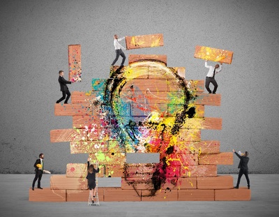 Zes mensen bouwen een muur uit kleurrijke bakstenen die samen een lamp voorstellen, als symbool voor een creatief idee dat geleidelijk vorm krijgt