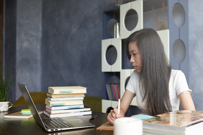Een jonge vrouw zit aan een bureau met stapels boeken en kijkt naar haar laptop, terwijl ze notities maakt op papier