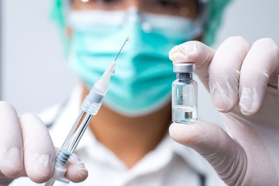 Ein Arzt mit Mund-Nasen-Schutz und medizinischen Handschuhen, der ein Fläschchen mit Impfstoff und eine Spritze hält