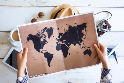 Valaki világtérképet tart a kezében, az asztalon mögötte egy tablet, egy csésze kávé, egy napszemüveg, egy kalap, egy fényképezőgép és egy repülőgépmodell látható, amelyek az utazást jelképezik